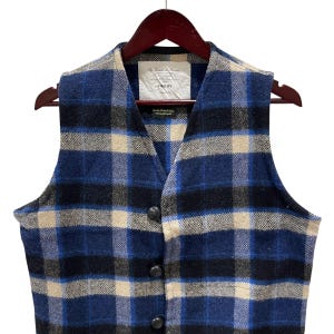 May include: A sleeveless vest featuring a blue, black, and cream plaid pattern. The garment has a V-neckline, a front button closure with three dark buttons, and a white interior label with the text Fidelity and other printed details.