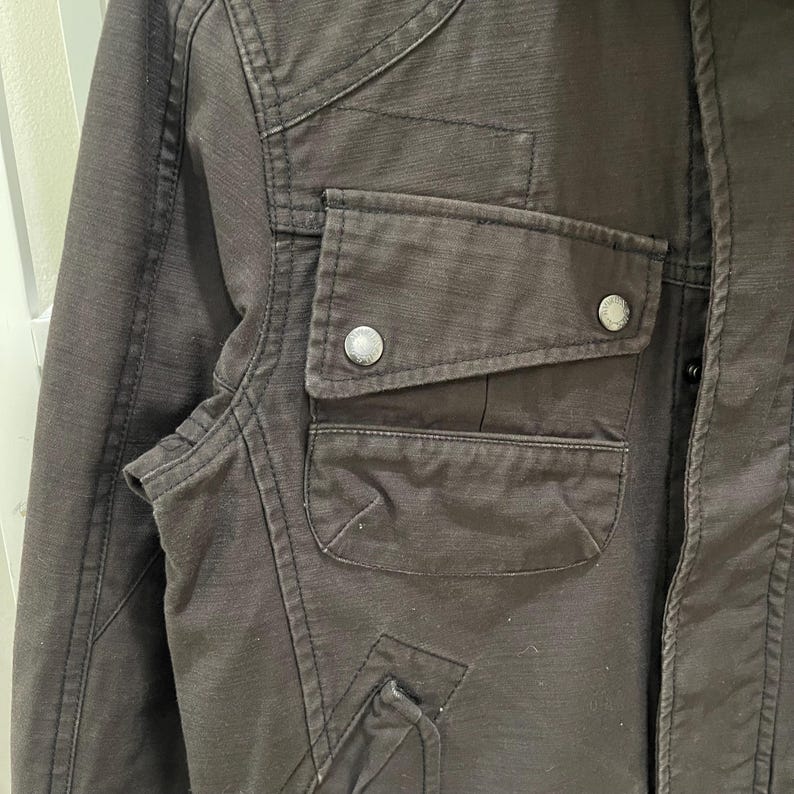 K&ouml;nnte beinhalten: Eine dunkelbraune Utility-Jacke aus strukturiertem Stoff. Das Kleidungsst&uuml;ck verf&uuml;gt &uuml;ber eine Brusttasche mit einer Klappe, die mit zwei silberfarbenen Metalldruckkn&ouml;pfen befestigt ist, verst&auml;rkte N&auml;hte und eine vertikale Knopfleiste.