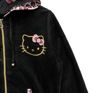 Pode incluir: Casaco de moletom preto de veludo com um design Hello Kitty dourado e um forro de leopardo rosa no capuz.