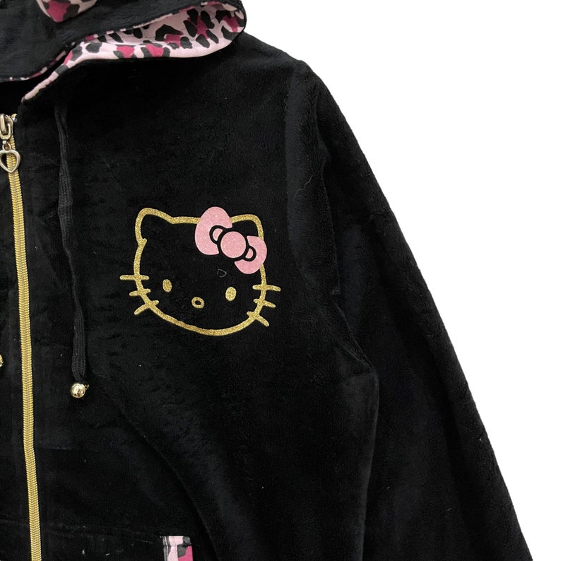 Pode incluir: Casaco de moletom preto de veludo com um design Hello Kitty dourado e um forro de leopardo rosa no capuz.