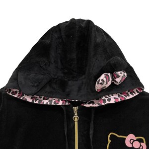 Pode incluir: Camisola de moletom com capuz preta com forro de estampa de leopardo rosa e preto. O capuz tem um forro de pel&uacute;cia preto e um la&ccedil;o de estampa de leopardo rosa e preto na parte superior. O z&iacute;per &eacute; dourado e tem um puxador em forma de cora&ccedil;&atilde;o. A parte frontal da camisola tem um gr&aacute;fico de Hello Kitty rosa.