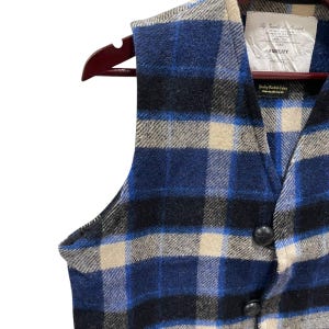 May include: A sleeveless vest featuring a blue, black, and beige plaid pattern. The garment has a V-neckline, two black buttons, and a white interior label reading By Gerald & Stewart FIDELITY, along with a smaller tag stating Quality Woolrich Fabric.