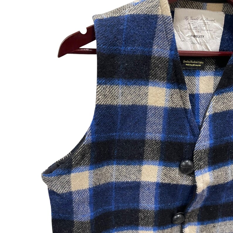 May include: A sleeveless vest featuring a blue, black, and beige plaid pattern. The garment has a V-neckline, two black buttons, and a white interior label reading By Gerald & Stewart FIDELITY, along with a smaller tag stating Quality Woolrich Fabric.