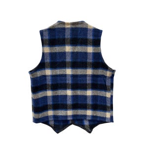 May include: A sleeveless vest featuring a plaid pattern in shades of blue, black, and cream. The garment has a high neckline, a straight back hem, and two pointed front panels that overlap at the center. The fabric has a textured, woven appearance.