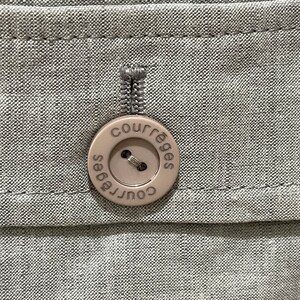 Puede incluir: Primer plano de una tela gris claro con un bot&oacute;n beige. El bot&oacute;n redondo tiene la palabra "courr&egrave;ges" impresa alrededor del borde. El bot&oacute;n est&aacute; cosido a la tela con hilo a juego.