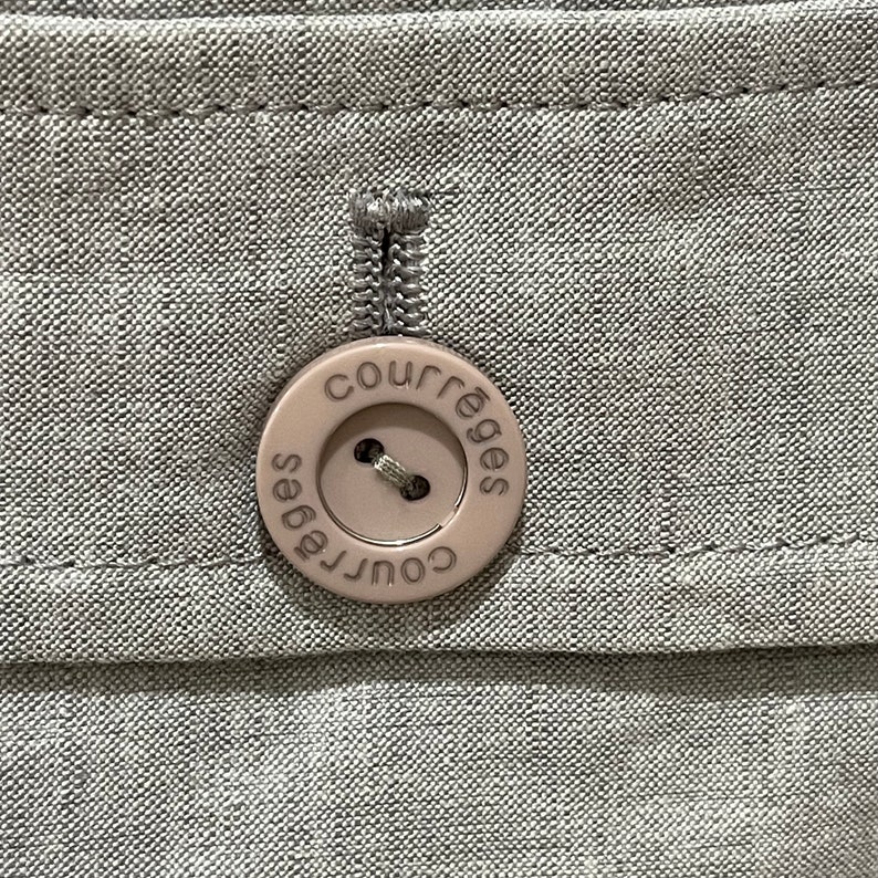 Puede incluir: Primer plano de una tela gris claro con un bot&oacute;n beige. El bot&oacute;n redondo tiene la palabra "courr&egrave;ges" impresa alrededor del borde. El bot&oacute;n est&aacute; cosido a la tela con hilo a juego.