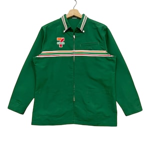 Puede incluir: Chaqueta de trabajo verde con cierre de cremallera y un logotipo de 7-Eleven en la parte delantera. La chaqueta tiene un diseño a rayas en el pecho y dos bolsillos en la parte delantera.