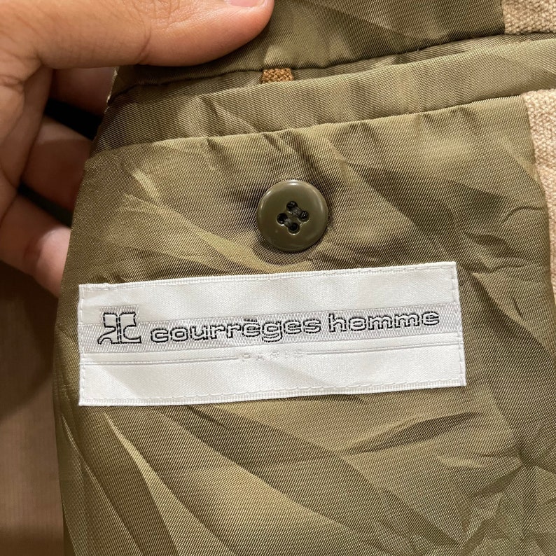 Puede incluir: Primer plano del interior de una blazer beige. El forro es verde oliva con un bot&oacute;n en el lado derecho. Una etiqueta blanca con el texto "courr&egrave;ges homme" est&aacute; cosida en el forro.