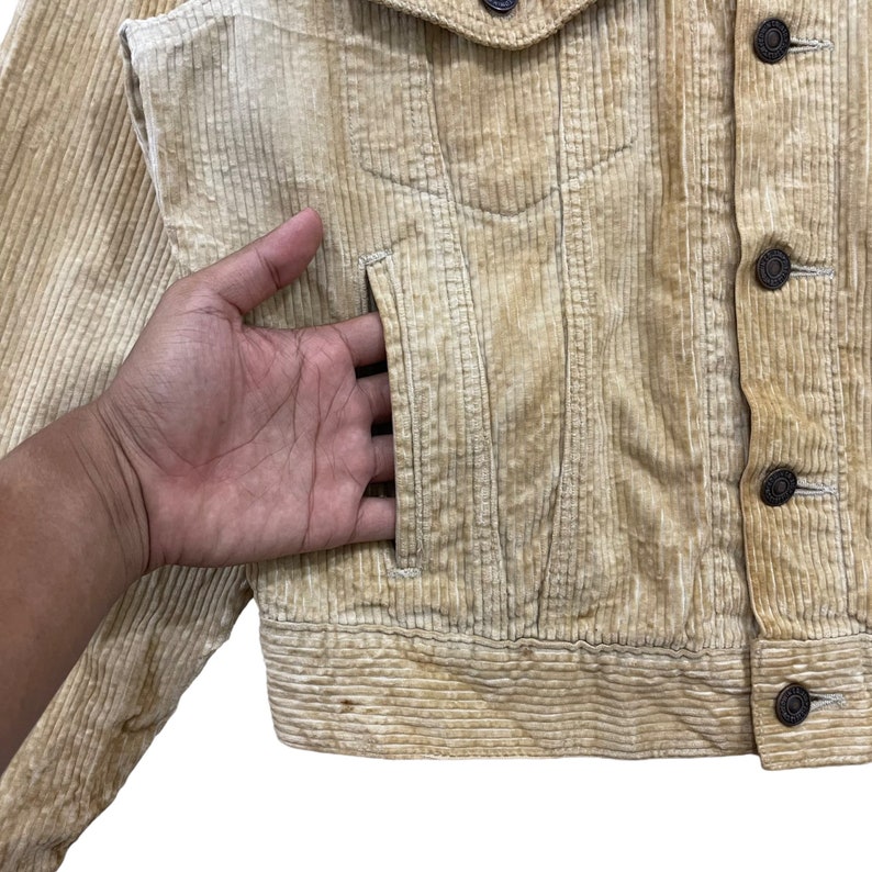 May include: A tan corduroy jacket with a button-up front and two chest pockets. The jacket has a classic design with a ribbed texture. The buttons are dark brown. The jacket is a neutral colour, suitable for various outfits.