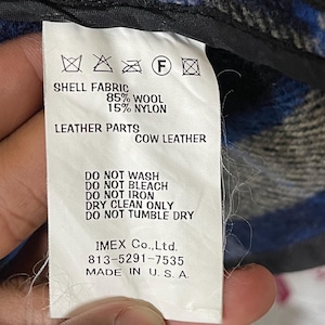 May include: A white garment care label featuring black text and symbols. It specifies shell fabric as 85% wool and 15% nylon, with cow leather parts. Care instructions include do not wash, bleach, iron, or tumble dry, and dry clean only. Made in U.S.A.