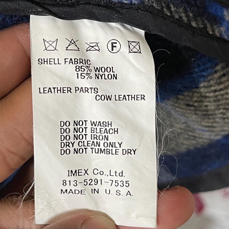 May include: A white garment care label featuring black text and symbols. It specifies shell fabric as 85% wool and 15% nylon, with cow leather parts. Care instructions include do not wash, bleach, iron, or tumble dry, and dry clean only. Made in U.S.A.
