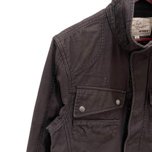 K&ouml;nnte beinhalten: Eine dunkelbraune Utility-Jacke mit hohem Kragen, verst&auml;rkten Schulterpartien und zwei Brusttaschen mit Druckknopfverschluss. Das Innenetikett zeigt den Text Avirex sowie weitere Herstellerangaben und die Gr&ouml;&szlig;enangabe M.
