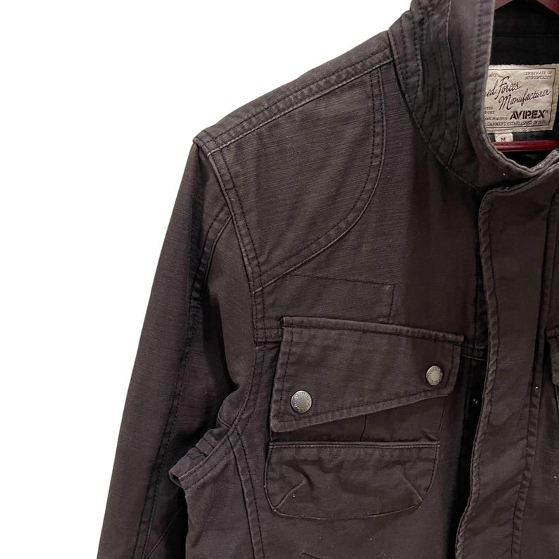 K&ouml;nnte beinhalten: Eine dunkelbraune Utility-Jacke mit hohem Kragen, verst&auml;rkten Schulterpartien und zwei Brusttaschen mit Druckknopfverschluss. Das Innenetikett zeigt den Text Avirex sowie weitere Herstellerangaben und die Gr&ouml;&szlig;enangabe M.