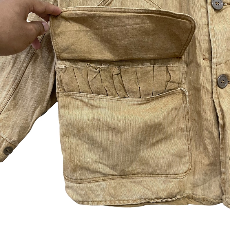 May include: A brown utility jacket with a large flap pocket on the front. The pocket has a pleated detail and a smaller pocket on the front.