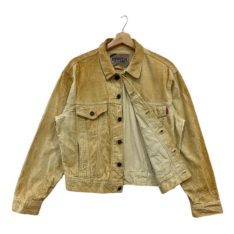 May include: A tan corduroy jacket with a button-up front, two chest pockets, and a small red tag. The jacket is hanging on a wooden hanger. The brand name "EDWIN" is visible on a tag.