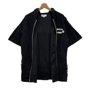 May include: Black hooded zip-up short-sleeved jacket with the text "KITTY MELODY" in white. The jacket has a white zip and is hanging on a wooden hanger. The jacket has a soft, plush texture.