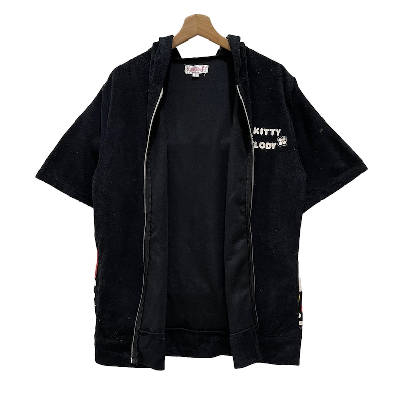 May include: Black hooded zip-up short-sleeved jacket with the text "KITTY MELODY" in white. The jacket has a white zip and is hanging on a wooden hanger. The jacket has a soft, plush texture.