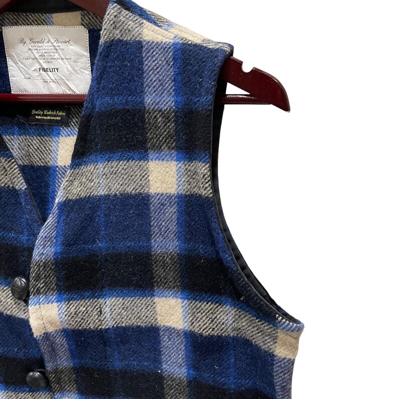 May include: A sleeveless vest featuring a blue, black, and cream plaid pattern. The garment has a V-neckline, dark buttons, and a white interior label with the text By Gerald & Stewart, Fidelity, and Quality Woolrich Fabric, Made in the USA.