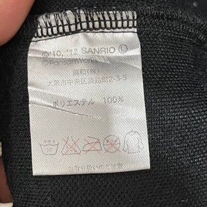 May include: A white label with black text and symbols, attached to a black fabric. The label includes the text "&copy;'10, '12 SANRIO", "&copy;PansonWorks", Japanese characters, and "ポリエステル 100%", along with washing instructions.
