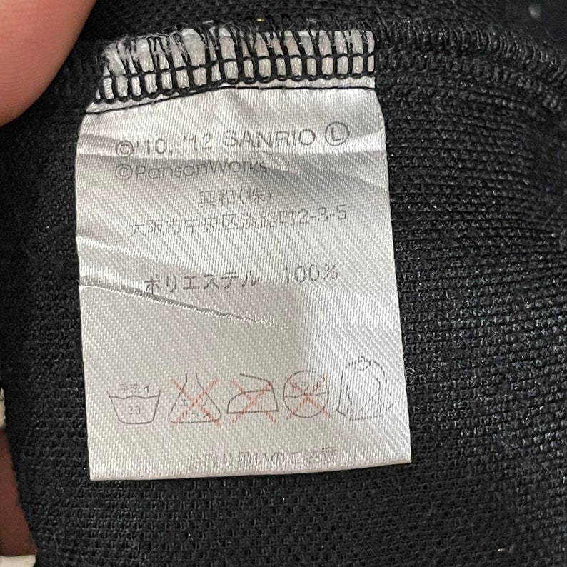 May include: A white label with black text and symbols, attached to a black fabric. The label includes the text "&copy;'10, '12 SANRIO", "&copy;PansonWorks", Japanese characters, and "ポリエステル 100%", along with washing instructions.