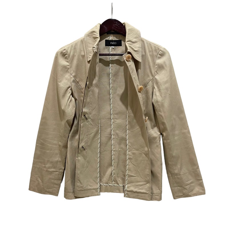 May include: Tan jacket with a collared neckline and button closures. The jacket has long sleeves and a tailored fit. The brand name "INED" is visible on the label. The jacket is hanging on a wooden hanger against a white background.