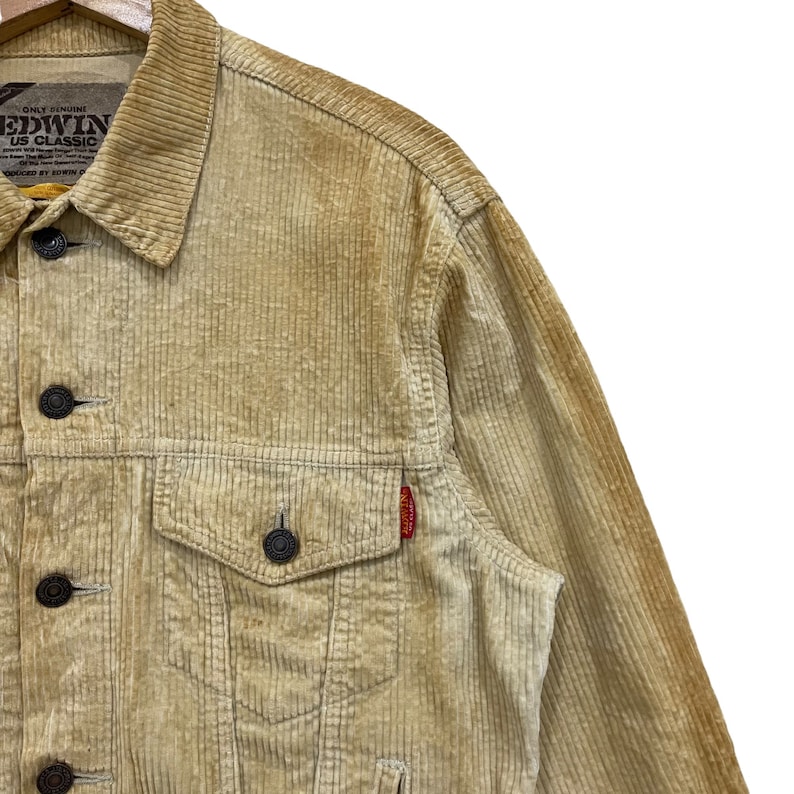 May include: A tan corduroy jacket with a button-down front, two chest pockets, and a small red tag. The jacket has a classic design with a collared neckline and dark buttons. The brand name "EDWIN US CLASSIC" is visible on the label.