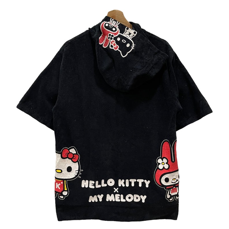 May include: Black short-sleeved hoodie with Hello Kitty and My Melody characters. The back features Hello Kitty and My Melody graphics, with the text "HELLO KITTY x MY MELODY". The hood has additional character designs.