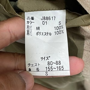 Puede incluir: Primer plano de una etiqueta de ropa con texto en japon&eacute;s. La etiqueta muestra el c&oacute;digo de producto J88617, el color 01 y la talla S. Enumera los materiales como 100% algod&oacute;n para la tela exterior y 100% poli&eacute;ster para el forro. Contorno de pecho 80-88 cm, altura 155-165 cm.