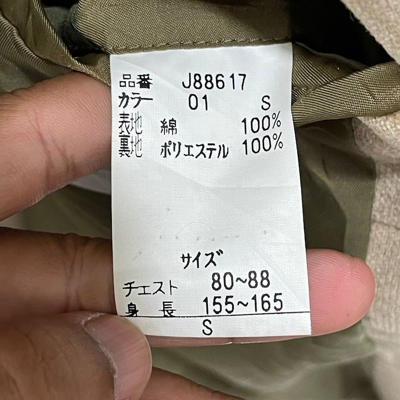 Puede incluir: Primer plano de una etiqueta de ropa con texto en japon&eacute;s. La etiqueta muestra el c&oacute;digo de producto J88617, el color 01 y la talla S. Enumera los materiales como 100% algod&oacute;n para la tela exterior y 100% poli&eacute;ster para el forro. Contorno de pecho 80-88 cm, altura 155-165 cm.