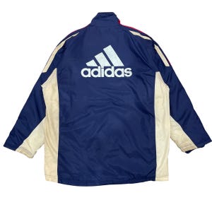 May include: A navy blue Adidas jacket with a large white Adidas logo on the back. The jacket has cream-colored side panels and red accents. The jacket has a full zipper and a high collar.
