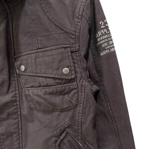K&ouml;nnte beinhalten: Eine dunkelbraune Jacke mit einer Brusttasche und zwei silberfarbenen Metalldruckkn&ouml;pfen. Auf dem &Auml;rmel befindet sich ein wei&szlig;er Aufdruck mit dem Text 23 SUPPLY ROCKWELL AERONAUTIC SER AME DIV CORONA ARMY AIR. Der Stoff weist eine sichtbare Webstruktur auf.