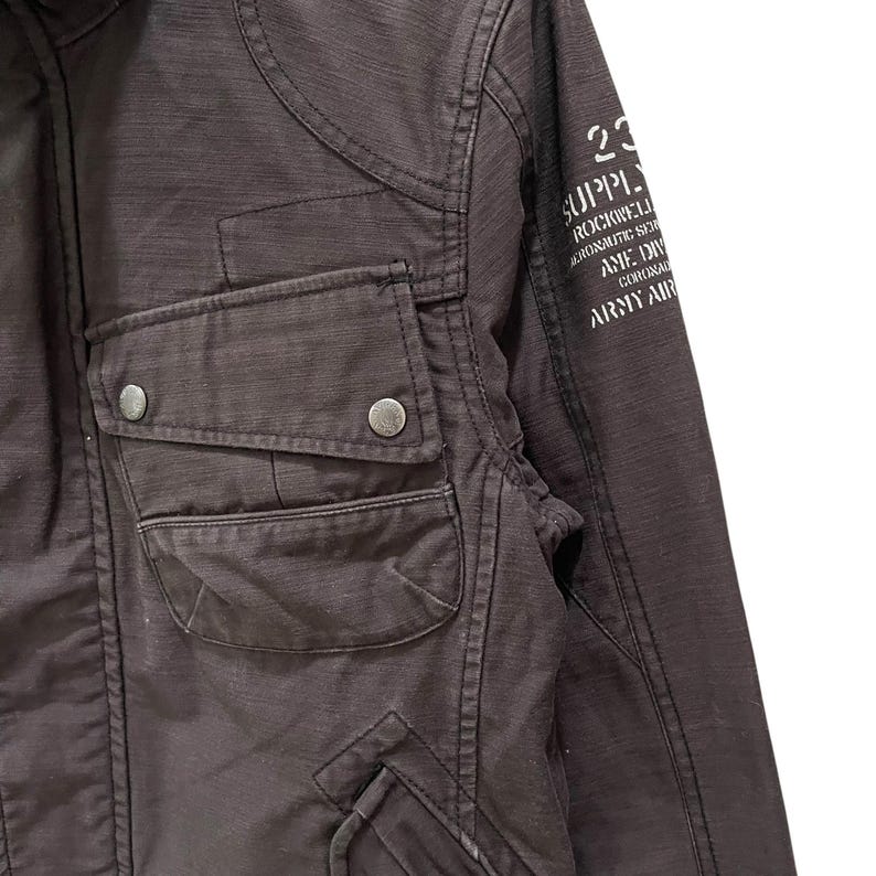 K&ouml;nnte beinhalten: Eine dunkelbraune Jacke mit einer Brusttasche und zwei silberfarbenen Metalldruckkn&ouml;pfen. Auf dem &Auml;rmel befindet sich ein wei&szlig;er Aufdruck mit dem Text 23 SUPPLY ROCKWELL AERONAUTIC SER AME DIV CORONA ARMY AIR. Der Stoff weist eine sichtbare Webstruktur auf.