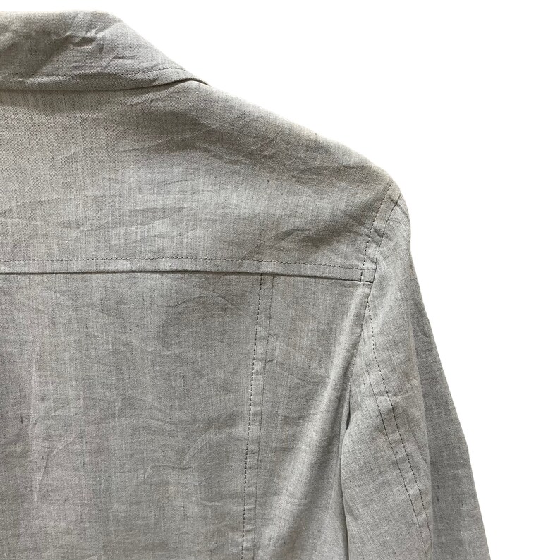 Puede incluir: Primer plano de una chaqueta gris claro. La chaqueta tiene un cuello y una costura en la espalda. La tela parece ser lino o un material ligero similar. Las costuras son visibles y a&ntilde;aden detalles a la prenda.