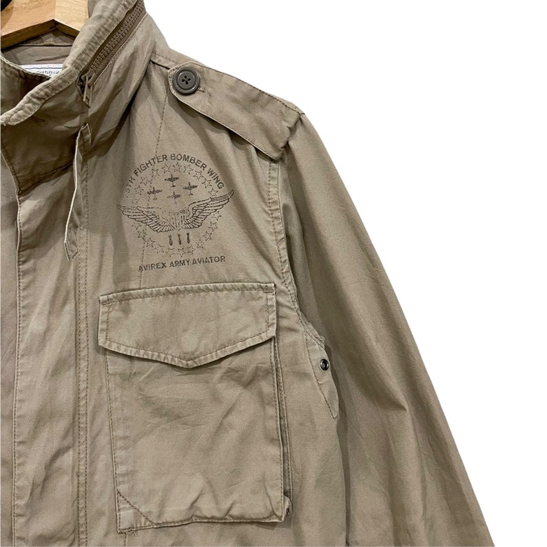 K&ouml;nnte beinhalten: Eine milit&auml;risch anmutende Jacke in Beige mit Rei&szlig;verschluss und mehreren Taschen. Die Jacke hat einen Knopf auf der Schulter und ein gedrucktes Emblem auf der Brust mit der Aufschrift "5th Fighter Bomber Wing" und "Avirex Army Aviator".