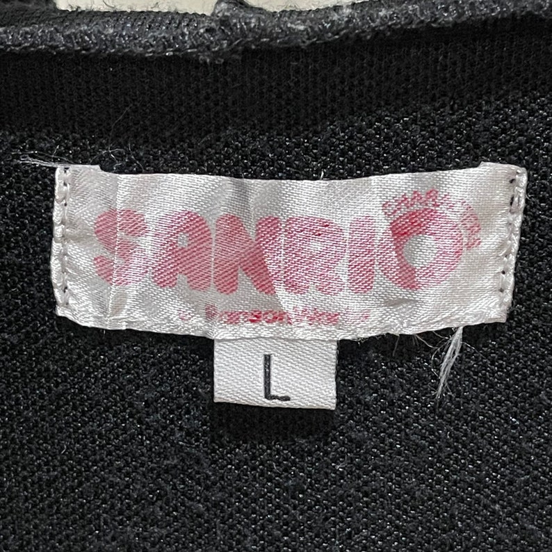 May include: A close-up of a black garment tag with the word "SANDRO" in pink letters. The tag also features a pink crown design and the size "L" in a white rectangle.