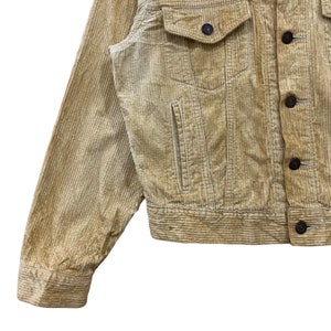 May include: A close-up shot of a tan corduroy jacket. The jacket features two chest pockets with button fastenings, a small pocket on the left, and a button-up front. The sleeves are long, and the fabric has a ribbed texture.