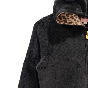 Peut inclure: Veste zipp&eacute;e en polaire noire avec une doublure en imprim&eacute; l&eacute;opard sur la capuche. La veste a une fermeture &eacute;clair dor&eacute;e et un logo dor&eacute; sur le tirette de la fermeture &eacute;clair.