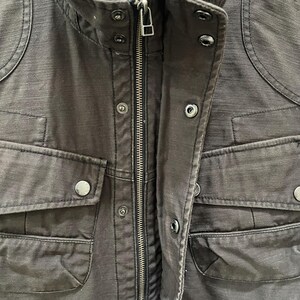 K&ouml;nnte beinhalten: Eine dunkel anthrazitfarbene Utility-Jacke mit zentralem Metallrei&szlig;verschluss, Stehkragen mit Druckkn&ouml;pfen und mehreren Vordertaschen mit Druckknopfklappen. Der Stoff weist eine deutlich sichtbare, texturierte Webstruktur auf.