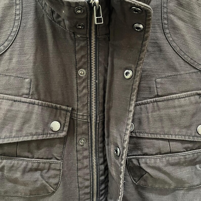 K&ouml;nnte beinhalten: Eine dunkel anthrazitfarbene Utility-Jacke mit zentralem Metallrei&szlig;verschluss, Stehkragen mit Druckkn&ouml;pfen und mehreren Vordertaschen mit Druckknopfklappen. Der Stoff weist eine deutlich sichtbare, texturierte Webstruktur auf.