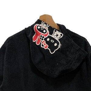 May include: Black hooded sweatshirt featuring a ninja and a cat graphic. The ninja is red with a white face, and the cat is white with black details. The sweatshirt is displayed on a wooden hanger.