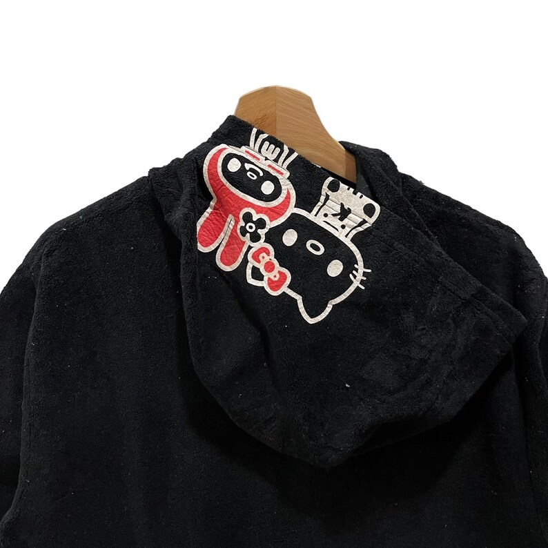 May include: Black hooded sweatshirt featuring a ninja and a cat graphic. The ninja is red with a white face, and the cat is white with black details. The sweatshirt is displayed on a wooden hanger.