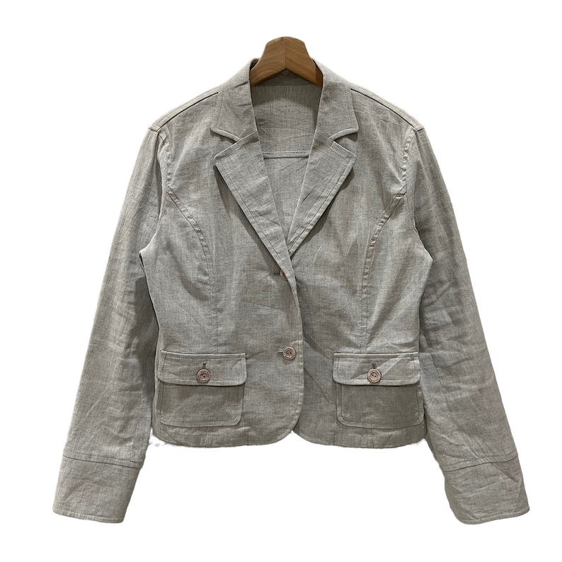 Puede incluir: Un blazer gris claro con cierre de un solo bot&oacute;n, dos bolsillos delanteros y mangas largas. El blazer est&aacute; hecho de una tela texturizada y se muestra en una percha de madera sobre un fondo blanco. El blazer tiene un dise&ntilde;o cl&aacute;sico y entallado.