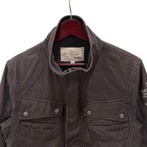 K&ouml;nnte beinhalten: Eine dunkelbraune Utility-Jacke mit Stehkragen, zwei Brusttaschen mit Druckknopfverschluss und einem rechteckigen Innenetikett mit der Aufschrift Armed Forces Manufacturer AVIREX. Die Jacke verf&uuml;gt &uuml;ber verst&auml;rkte Schulterpartien und lange &Auml;rmel.