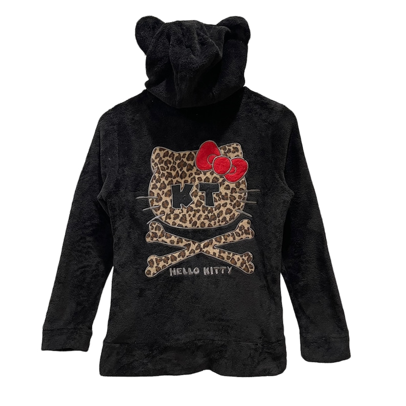 Peut inclure: Sweat-shirt &agrave; capuche noir avec un motif Hello Kitty imprim&eacute; l&eacute;opard avec un cr&acirc;ne et des os crois&eacute;s. Le texte "Hello Kitty" est imprim&eacute; sous le motif.