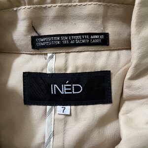 May include: Close-up of a beige jacket's interior, showcasing the brand label "IN&Eacute;D" on a black tag. Below, a white tag displays the number "7". Above, a black tag reads "COMPOSITION SUR ETIQUETTE ANNEXE COMPOSITION: SEE ATTACHED LABEL".
