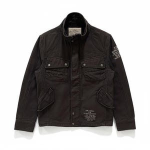 K&ouml;nnte beinhalten: Eine dunkelbraune Utility-Jacke mit Stehkragen, vier aufgesetzten Pattentaschen mit Druckkn&ouml;pfen und einer zentralen Druckknopfleiste. Auf dem linken &Auml;rmel und dem unteren Vorderteil befindet sich wei&szlig;er Text, darunter "23 SUPPLY" und "AIR CORPS".