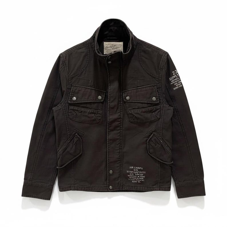 K&ouml;nnte beinhalten: Eine dunkelbraune Utility-Jacke mit Stehkragen, vier aufgesetzten Pattentaschen mit Druckkn&ouml;pfen und einer zentralen Druckknopfleiste. Auf dem linken &Auml;rmel und dem unteren Vorderteil befindet sich wei&szlig;er Text, darunter "23 SUPPLY" und "AIR CORPS".