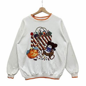 Könnte beinhalten: Weißes Sweatshirt mit einem Halloween-Motiv. Das Design zeigt einen Kürbis, einen Teddybären und eine Einkaufstasche voller Süßigkeiten und Geschenke. Der Text "Trick or Treat" ist ebenfalls sichtbar. Der Kragen und die Manschetten haben einen orangefarbenen Besatz.