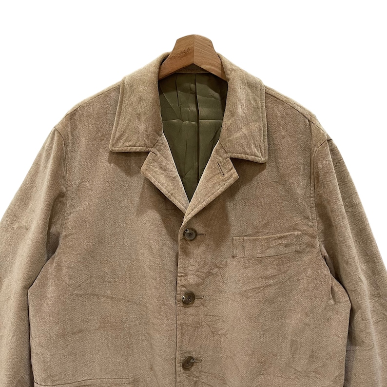 Puede incluir: Un blazer color beige con solapas y tres botones marrones. La chaqueta tiene un bolsillo y est&aacute; colgada de una percha de madera. El forro interior es verde oliva.