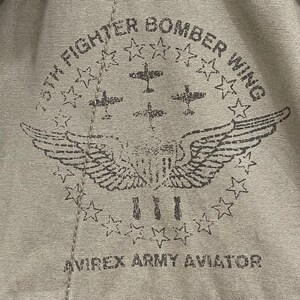 K&ouml;nnte beinhalten: Nahaufnahme eines beigefarbenen Stoffes mit einem Used-Print. Der Druck zeigt ein kreisf&ouml;rmiges Design mit Sternen, Fl&uuml;geln und dem Text "8th Fighter Bomber Wing" und "Avirex Army Aviator".